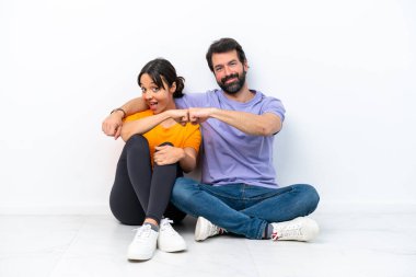 Young caucasian couple sitting on the floor isolated on white background bumping fists