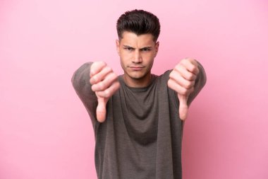 Pembe arka planda izole edilmiş genç beyaz adam baş parmağını iki eliyle aşağı gösteriyor.