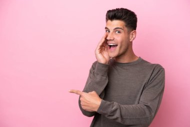 Pembe arka planda izole edilmiş genç beyaz bir adam bir ürün sunmak ve bir şeyler fısıldamak için kenarı işaret ediyor.