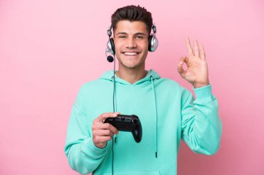 Pembe arka planda izole edilmiş bilgisayar oyunu kontrolörüyle oynayan yakışıklı beyaz bir adam parmaklarıyla tamam işareti gösteriyor.