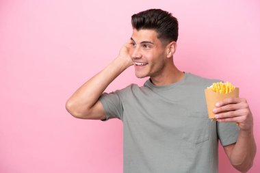 Pembe arka planda izole edilmiş patates kızartması tutan genç beyaz bir adam kulağına elini sürerek bir şeyler dinliyor.