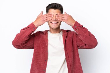 Beyaz arka planda izole edilmiş beyaz tenli genç bir adam. Gözleri elleriyle kaplıyor ve gülümsüyor.
