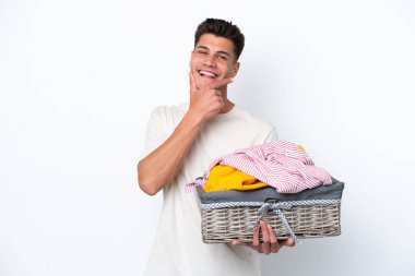 Beyaz arka planda çamaşır sepetini tutan genç beyaz adam mutlu ve gülümsüyor.