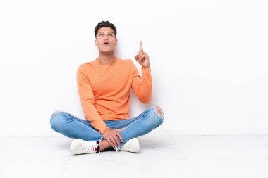 Yerde oturan genç adam beyaz arka planda izole edilmiş yukarı bakıyor ve şaşırıyor.
