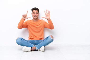 Yerde oturan genç adam beyaz arka planda izole edilmiş yediyi parmaklarıyla sayıyor.