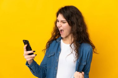 Sarı arka planda izole edilmiş genç Rus kız cep telefonu kullanıyor ve zafer jesti yapıyor.