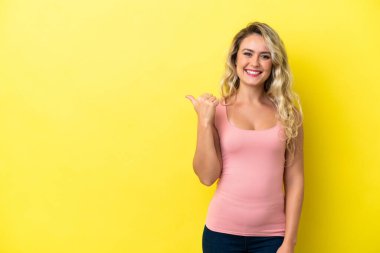Sarı arka planda izole edilmiş genç Brezilyalı kadın bir ürün sunmak için kenarı işaret ediyor.