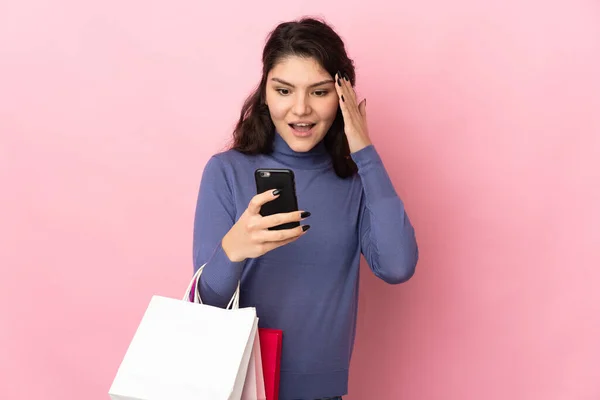 十代のロシアの女の子はピンクの背景に隔離されたショッピングバッグを保持し 友人に彼女の携帯電話でメッセージを書く — ストック写真