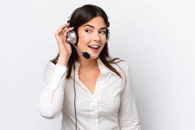 Beyaz arka planda izole edilmiş kulaklıkla çalışan beyaz pazarlamacı kadın elini kulağına koyarak bir şeyler dinliyor.