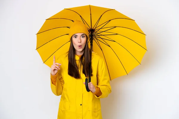 指を持ち上げながら解決策を実現しようと白い背景に隔離されたレインプルーフコートと傘を持つ若いブラジル人女性 — ストック写真