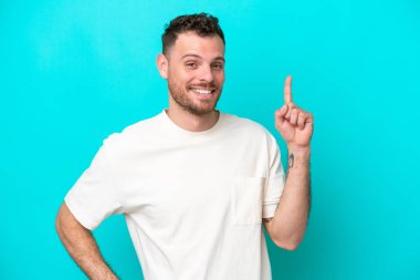 Genç Brezilyalı bir adam mavi arka planda izole edilmiş ve en iyinin işareti olarak parmağını kaldırıyor.