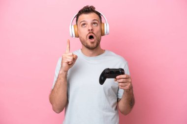 Young Brazilian man playing with video game controller isolated on pink background thinking an idea pointing the finger up