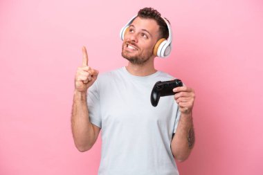 Young Brazilian man playing with video game controller isolated on pink background pointing up a great idea