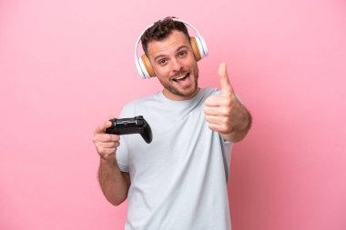 Young Brazilian man playing with video game controller isolated on pink background with thumbs up because something good has happened