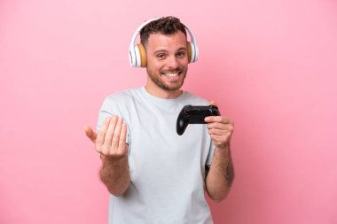 Young Brazilian man playing with video game controller isolated on pink background inviting to come with hand. Happy that you came