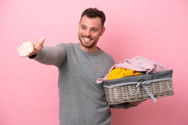Genç Brezilyalı bir adam pembe arka planda izole edilmiş bir çamaşır sepeti tutuyor ve baş parmak işareti yapıyor.