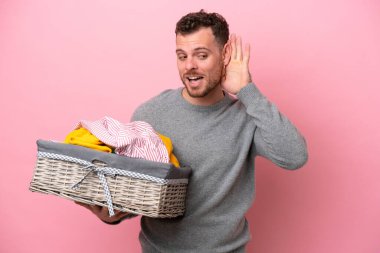 Genç Brezilyalı bir adam pembe arka planda izole edilmiş bir çamaşır sepeti tutuyor. Kulağına elini sürerek bir şeyler dinliyor.