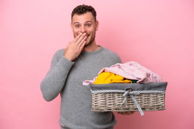 Genç Brezilyalı bir adam pembe arka planda izole edilmiş bir çamaşır sepeti tutuyor mutlu ve ağzı eliyle kaplayan gülümsüyor.