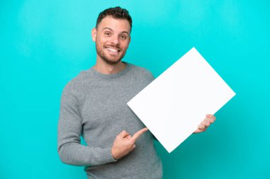 Genç Brezilyalı bir adam elinde boş bir levha tutuyor mavi arka planda izole edilmiş mutlu bir ifadeyle boş bir levha tutuyor ve onu işaret ediyor.