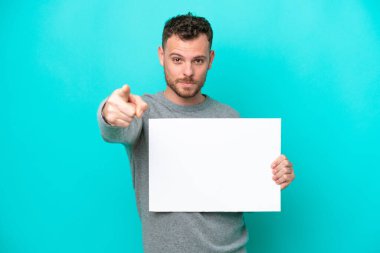 Elinde boş bir levha tutan genç Brezilyalı adam mavi arka planda izole edilmiş boş bir levha tutuyor ve ön tarafı işaret ediyor.