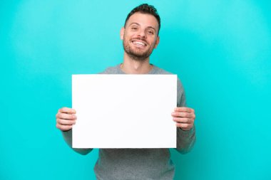 Elinde boş bir levha tutan genç Brezilyalı adam mavi arka planda izole edilmiş mutlu bir ifadeyle boş bir pankart tutuyor.