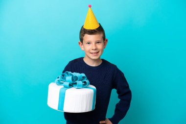 Küçük çocuk mavi arka planda izole edilmiş bir doğum günü pastası tutuyor kolları kalçasında ve gülümsüyor