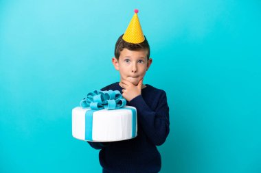 Küçük çocuk mavi arka planda izole edilmiş doğum günü pastası tutuyor. Şüpheleri ve düşünceleri var.