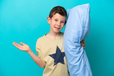 Mavi arka planda izole edilmiş pijamalı küçük çocuk, davet edilmek için ellerini yana uzatıyor.