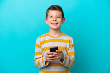 Küçük çocuk mavi arka planda izole edilmiş kameraya bakıyor ve cep telefonunu kullanarak gülümsüyor.