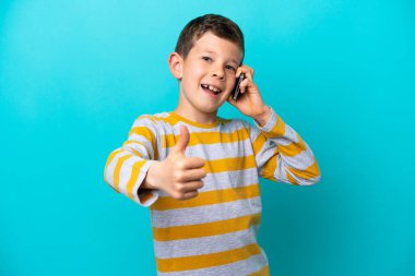 Küçük çocuk mavi arka planda izole edilmiş. Başparmaklarını kaldırırken cep telefonuyla sohbet ediyor.