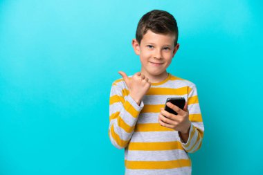 Küçük çocuk mavi arka planda cep telefonuyla izole edilmiş ve yan tarafı işaret ediyor.