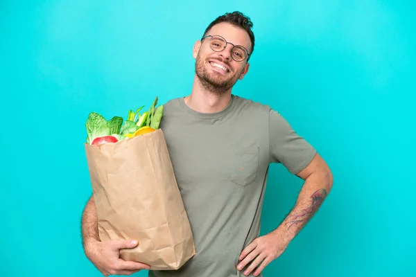 Young Brazilian man holding a grocery shopping bag isolated on blue background posing with arms at hip and smiling