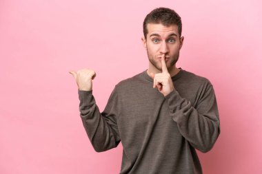Pembe arka planda izole edilmiş genç beyaz bir adam. Yan tarafı işaret ediyor ve sessizlik hareketi yapıyor.