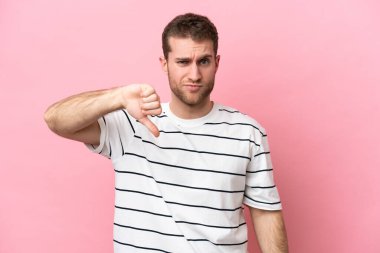 Pembe arka planda izole edilmiş genç beyaz adam olumsuz ifadeyle baş parmağını aşağı gösteriyor.