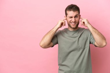 Pembe arka planda izole edilmiş genç beyaz adam hayal kırıklığına uğramış ve kulaklarını kapatmış.