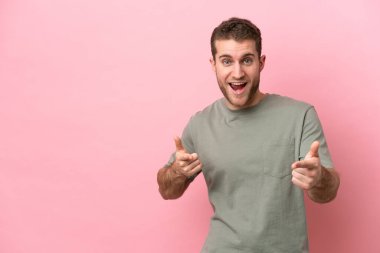 Pembe arka planda izole edilmiş genç beyaz adam ön tarafı işaret ediyor ve gülümsüyor.