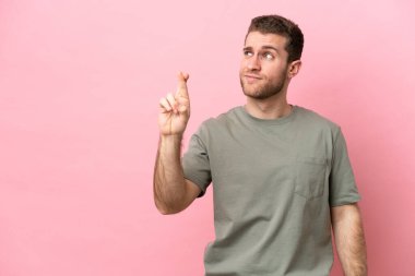 Pembe arka planda izole edilmiş genç beyaz adam parmaklarını çapraz yapıp en iyisini diliyor.