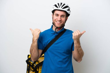 Beyaz arka planda izole edilmiş termal sırt çantalı beyaz tenli genç beyaz bir adam. Baş parmakları havada ve gülümsüyor.