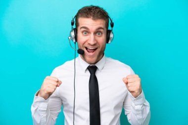 Tele pazarlamacı beyaz adam mavi arka planda izole edilmiş bir kulaklıkla çalışarak galibiyeti kutluyor.