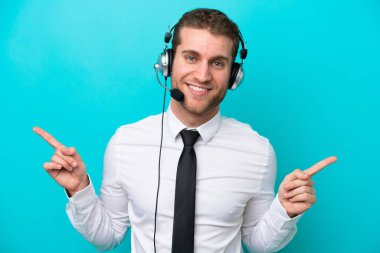 Tele pazarlamacı beyaz adam mavi arka planda izole edilmiş bir kulaklıkla çalışıyor. Parmak uçlarını işaret ediyor ve mutlu.
