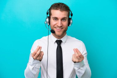 Tele pazarlamacı beyaz adam mavi arka planda izole edilmiş bir kulaklıkla çalışıyor. Para hareketi yapıyor.