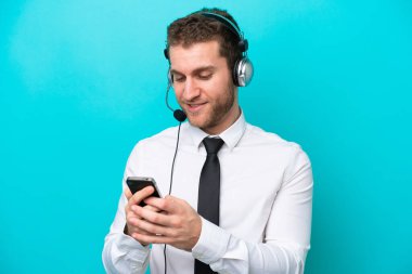 Tele pazarlamacı beyaz adam mavi arka planda izole edilmiş bir kulaklıkla çalışıyor cep telefonuyla bir mesaj gönderiyor.