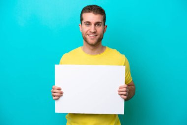 Young caucasian man isolated on blue background holding an empty placard with happy expression