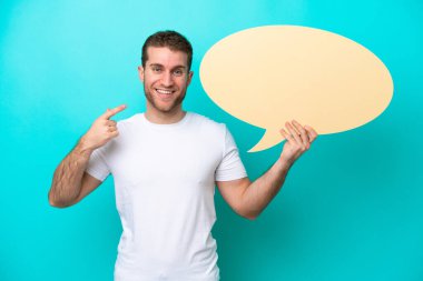 Young caucasian man isolated on blue background holding an empty speech bubble and pointing it