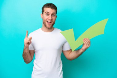 Kafkasyalı genç adam mavi arka planda izole edilmiş. Elinde bir kontrol simgesi var ve yukarı bakıyor.