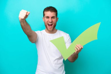 Mavi arka planda izole edilmiş genç beyaz bir adam elinde bir çek ikonu tutuyor ve zaferi kutluyor.