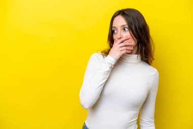 Sarı arka planda izole edilmiş genç beyaz kadın yan tarafa bakarken sürpriz jest yapıyor.