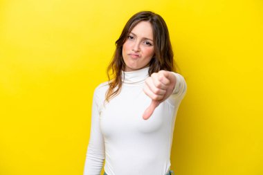 Sarı arka planda izole edilmiş genç beyaz kadın olumsuz ifadeyle baş parmağını aşağı gösteriyor.
