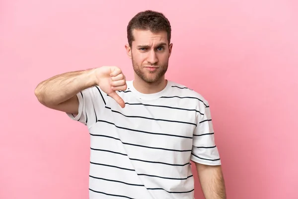 ピンクの背景に孤立した若い白人男性は 否定的な表現で親指を下に示す — ストック写真