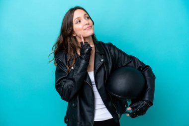 Motosiklet kasklı genç beyaz kadın mavi arka planda izole edilmiş yukarı bakarken bir fikir düşünüyor.